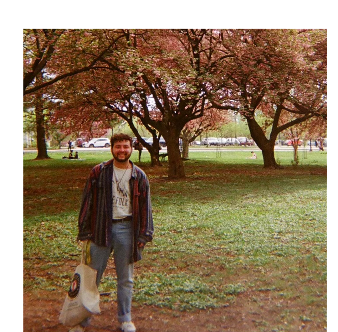 A grainy film camera produced image of Eli standing in front of cherry blossom trees in a park